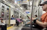 Passengers seated on a quiet Japanese commuter train with purple bench seats, hanging grab handles and colourful overhead adverts.